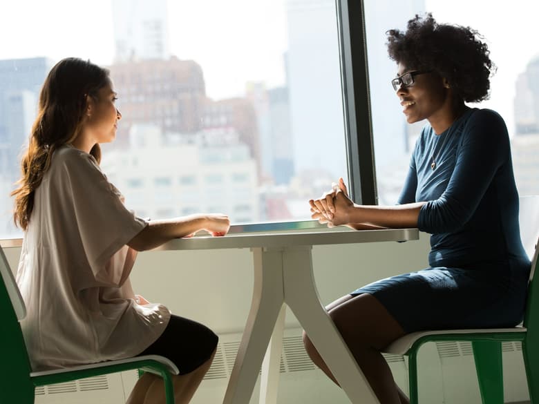 Dos profesionales conversan sentadas a una mesa junto a un ventanal, con edificios de la ciudad al fondo.